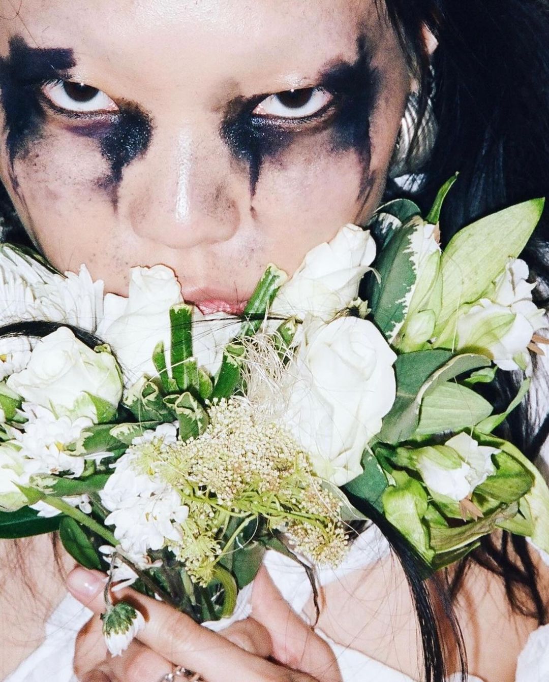 Close-up portrait of a person with black eye makeup holding a bouquet of white and green flowers, with a fair-skinned face and dark hair.