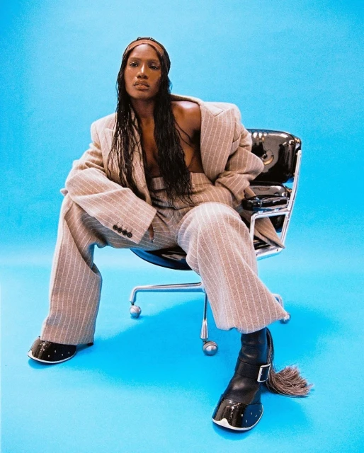 Fashionable woman in pinstripe suit sits in office chair against a blue backdrop, showcasing a blend of professional and edgy style.
