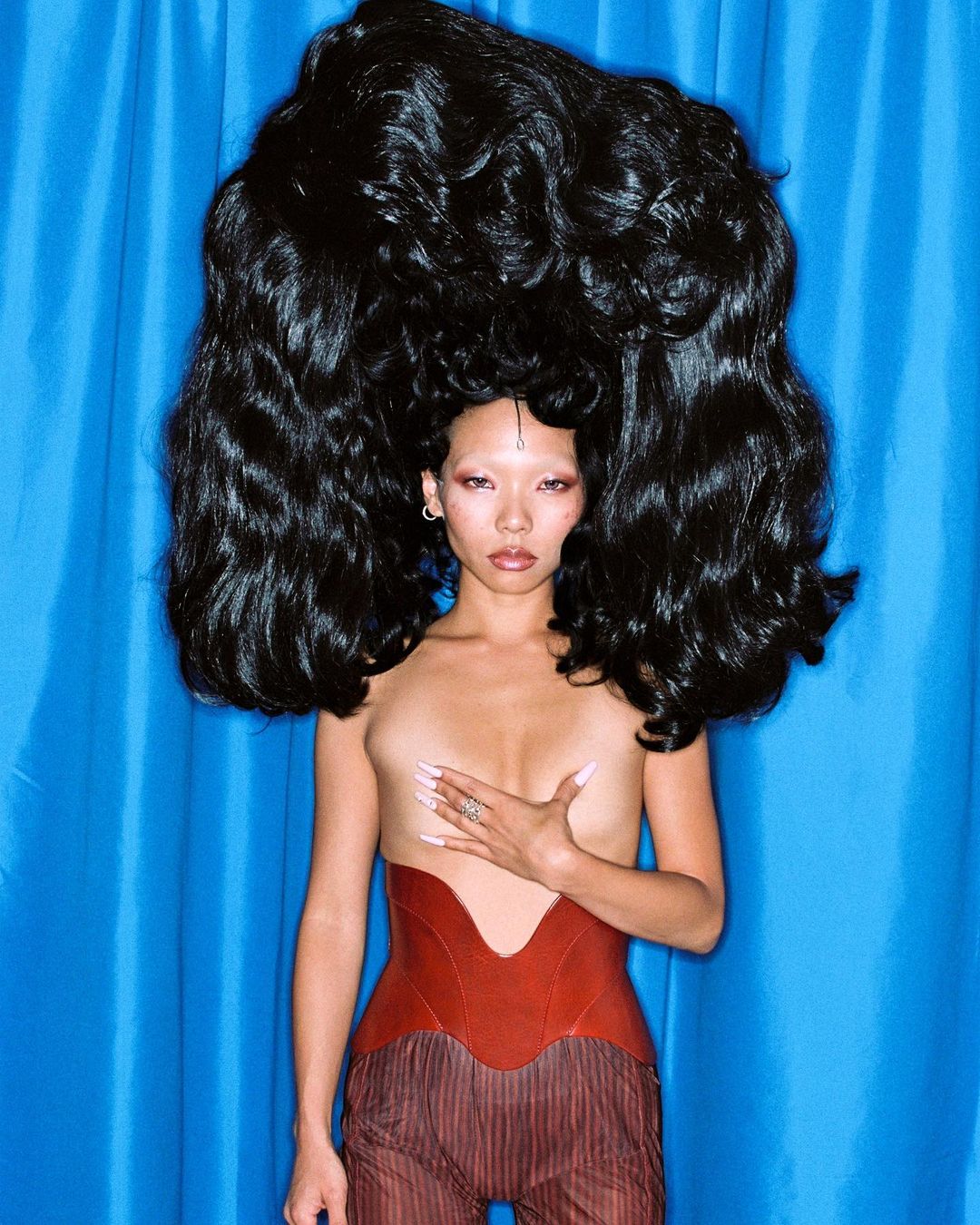 Striking studio portrait of a topless Asian woman with large black hair, wearing a red corset, against a bright blue backdrop.