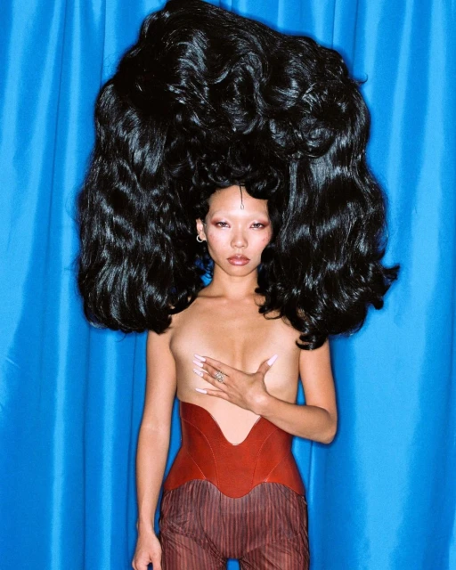 Striking studio portrait of a topless Asian woman with large black hair, wearing a red corset, against a bright blue backdrop.