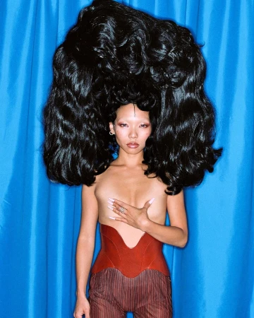 Striking studio portrait of a topless Asian woman with large black hair, wearing a red corset, against a bright blue backdrop.