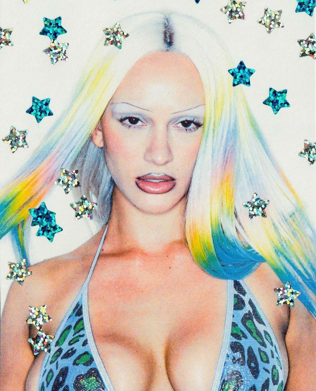 Close-up studio portrait of a woman with platinum and rainbow-colored hair, wearing a bikini top, against a white background with star decals.