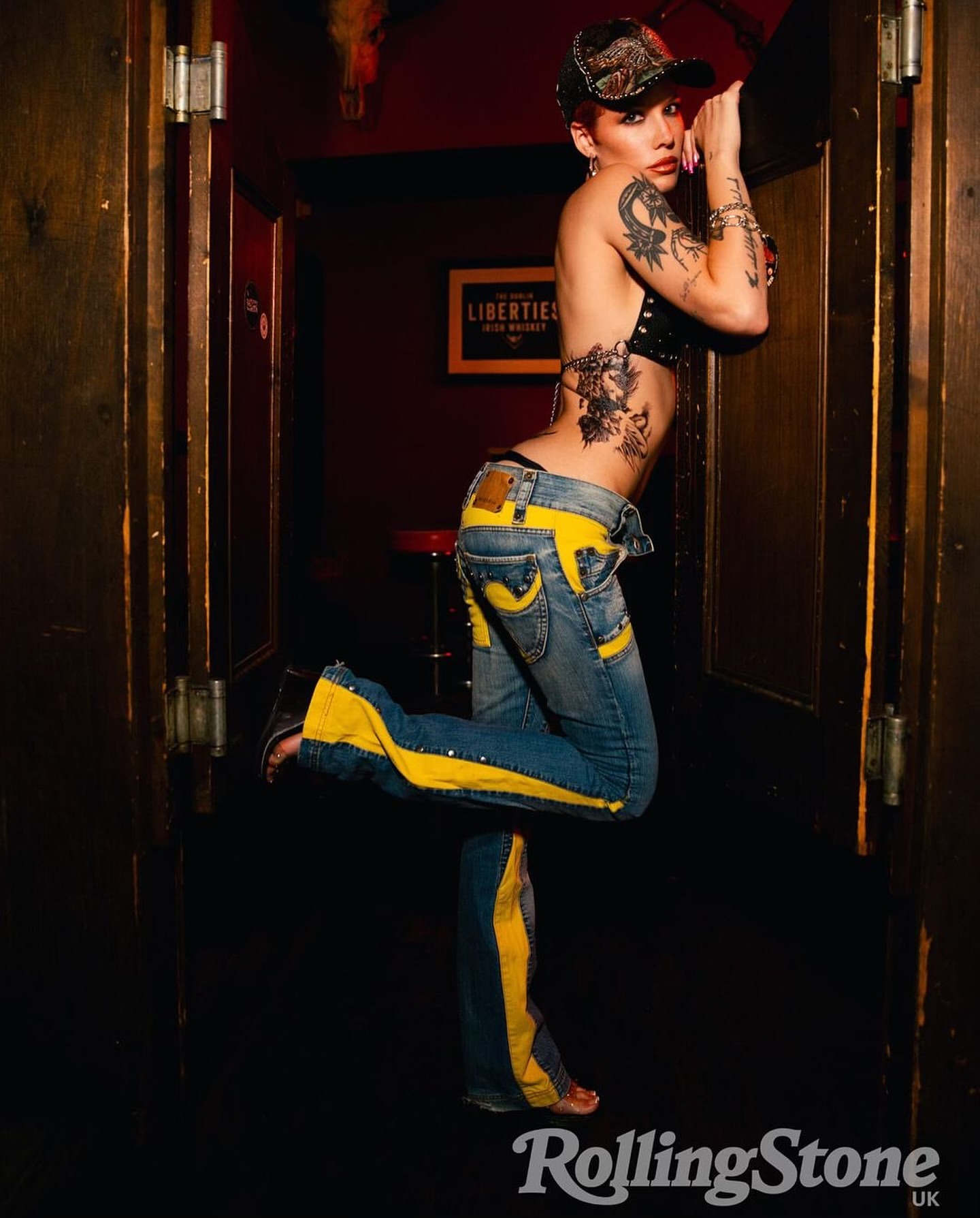 Stylish woman poses indoors, showcasing tattoos and fashion-forward jeans with yellow stripes in a dimly lit environment with Rolling Stone logo.