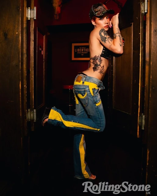 Stylish woman poses indoors, showcasing tattoos and fashion-forward jeans with yellow stripes in a dimly lit environment with Rolling Stone logo.
