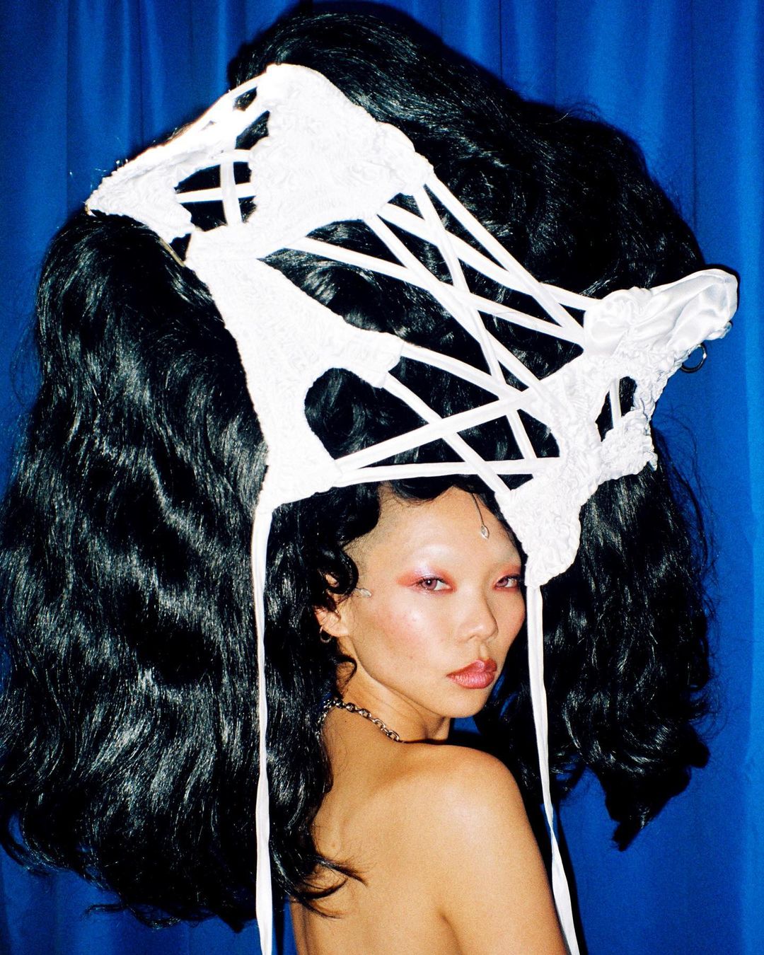 Striking close-up of a woman with creative dark hair and a white geometric headpiece against a vibrant blue backdrop in a studio setting.