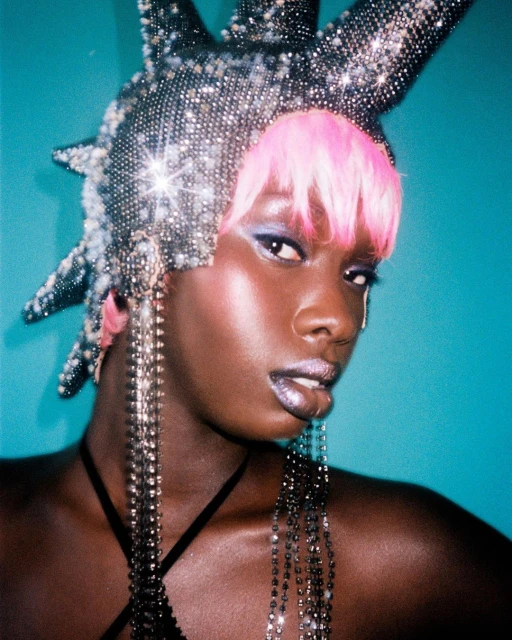 Close-up of a woman with dark skin wearing a spiked, sparkling headpiece and jeweled earrings against a turquoise background.