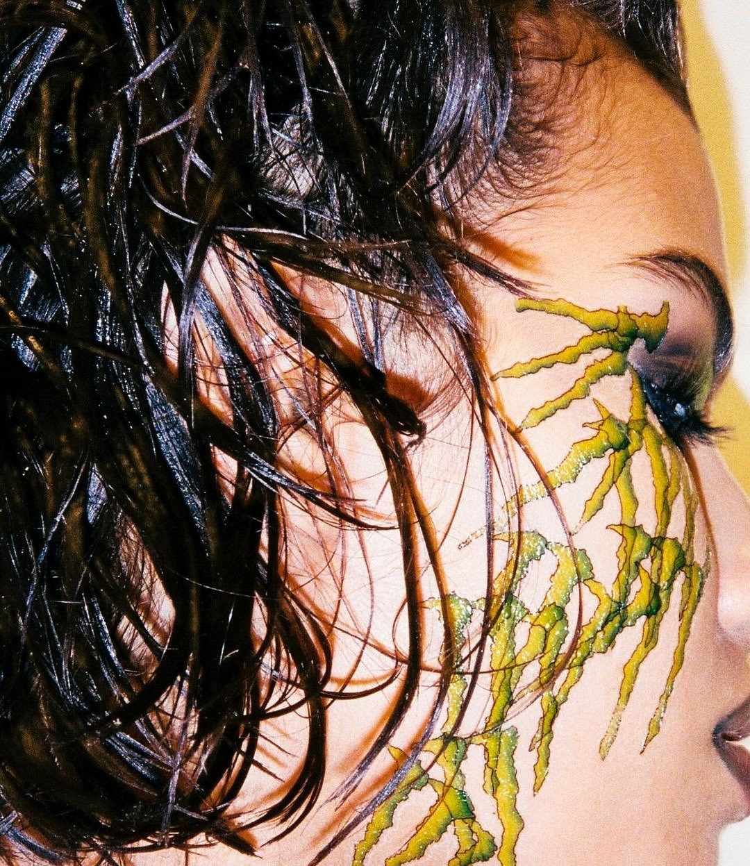 Close-up portrait of an adult female with dramatic green and black face paint, dark, slicked-back hair, and subtle makeup on warm-toned skin.
