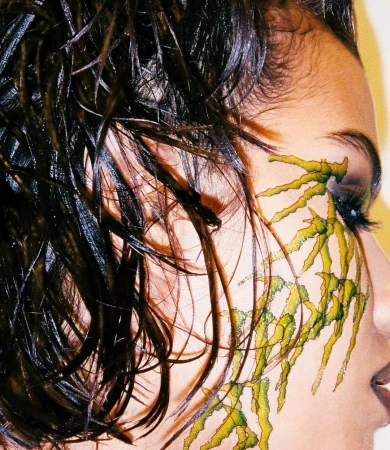 Close-up portrait of an adult female with dramatic green and black face paint, dark, slicked-back hair, and subtle makeup on warm-toned skin.