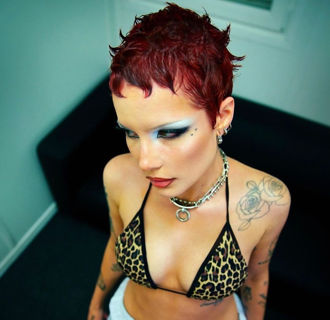 Adult female with red hair and heavy eye makeup wears a leopard-print bikini top and silver chain necklace looking down in an indoor shot.