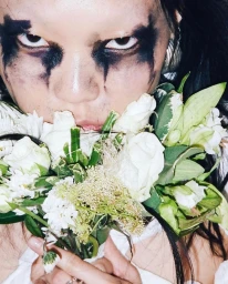 Close-up portrait of a person with heavy dark smeared eye makeup holding a bouquet of white roses and green foliage.