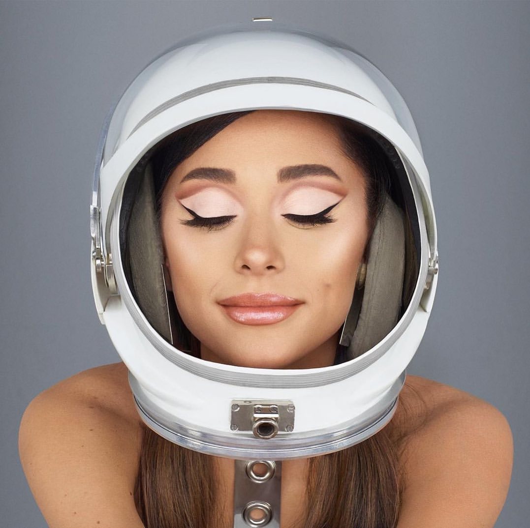 Adult woman with closed eyes wearing a white and silver astronaut helmet against a grey backdrop in a studio headshot.
