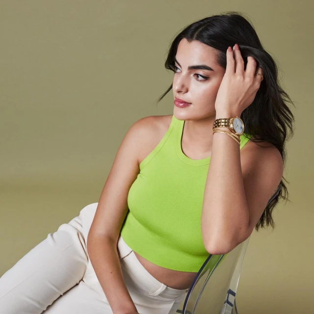 A brunette woman with tan skin and a lime-green tank top poses indoors with a gold watch and bracelets, seated in a clear chair.