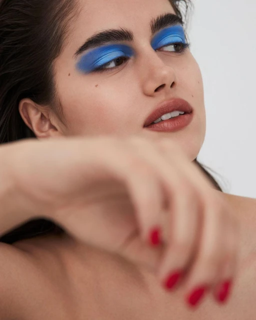 Woman with bold blue eyeshadow poses in a beauty shot against a white backdrop, showcasing makeup artistry and contemporary style.
