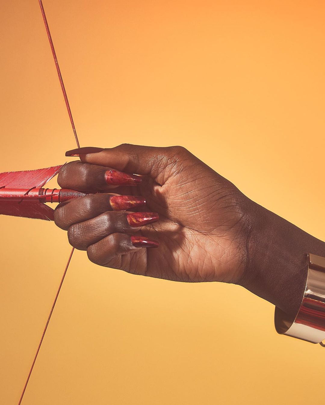 A dark-skinned hand with long red nails holds a red bow and arrow against an orange backdrop, emphasizing beauty and strength.