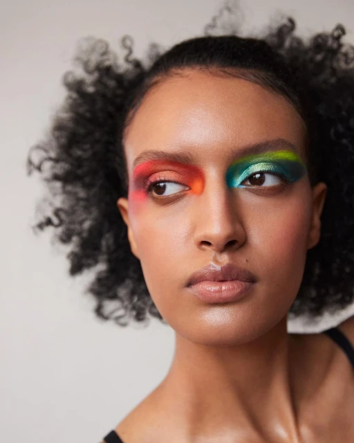 Close-up portrait of a Black female model with afro hair and creative colorful eye makeup on a neutral background, shot in studio lighting.