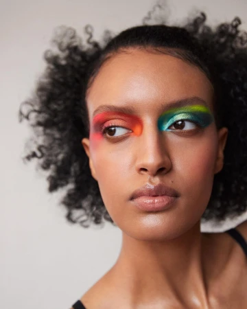Close-up portrait of a Black female model with afro hair and creative colorful eye makeup on a neutral background, shot in studio lighting.