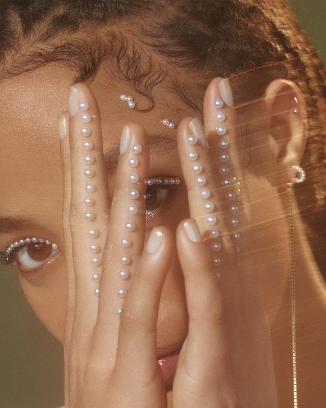 Close-up beauty shot of a woman partially veiled by her hands adorned with pearl embellishments, complemented by matching pearl eye makeup.