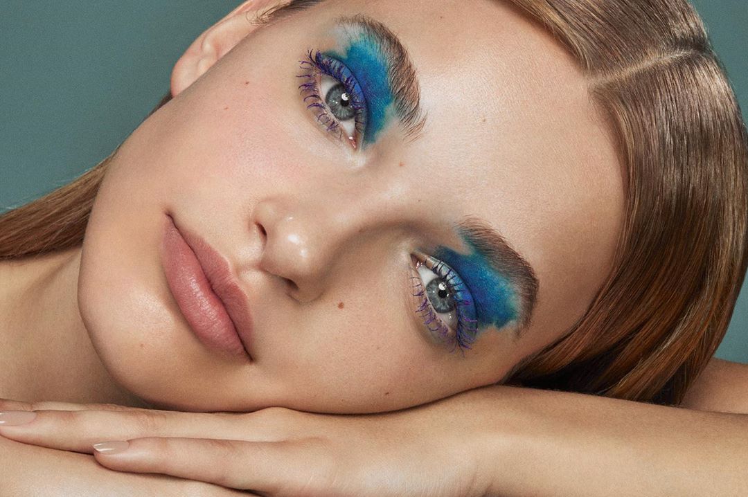 Close-up portrait of a woman with vibrant blue and purple makeup around her eyes, light skin, and light brown hair, set against a teal background.