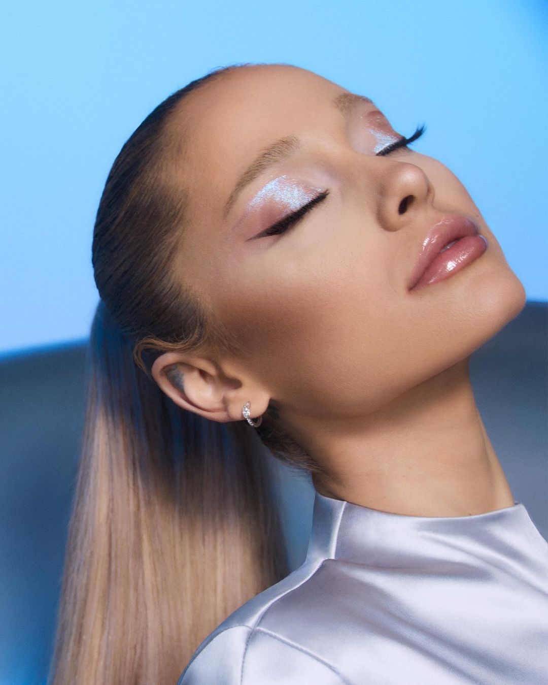 Close-up of a woman with shimmery makeup, closed eyes, and elegant ponytail, against a soft blue gradient background. Beauty shot.