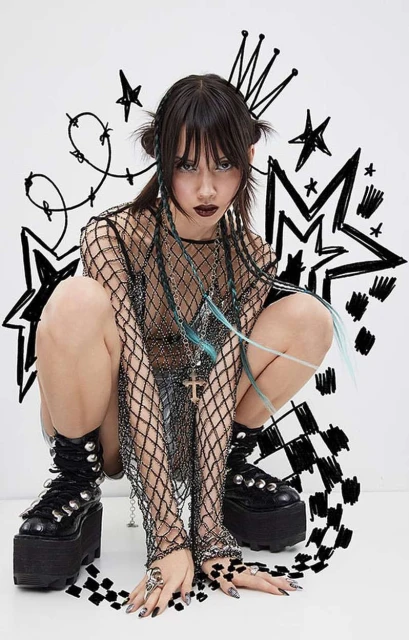 A young adult woman with dark hair and dark makeup crouches, wearing a black fishnet top, platform boots, and rings against a white background.