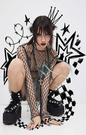 A young adult woman with dark hair and dark makeup crouches, wearing a black fishnet top, platform boots, and rings against a white background.