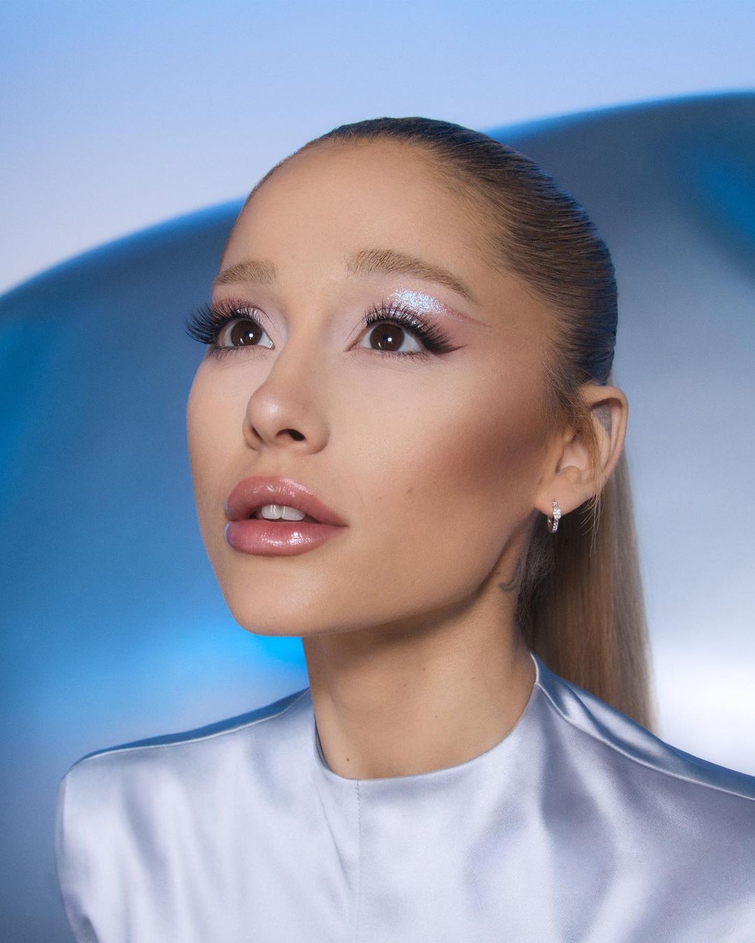Close-up portrait of an adult woman with light skin, light brown hair in a high ponytail, and a glossy lip, against a light blue backdrop.