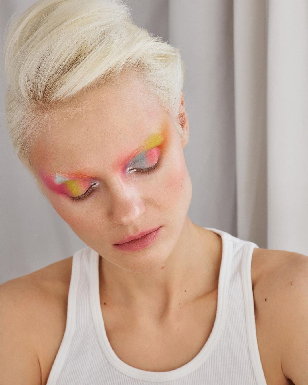 Close-up shot of a fair-skinned adult female model with short platinum blonde hair and vibrant, multi-colored eyeshadow makeup in a studio setting.