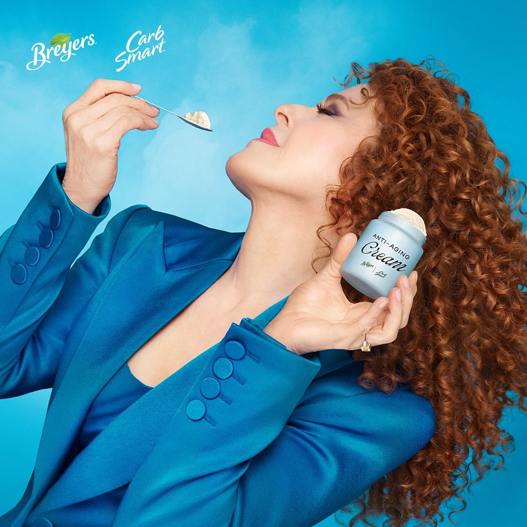 Bernadette Peters eats ice cream from a spoon, holding a container labeled "Anti-Aging Cream," in a light blue setting.