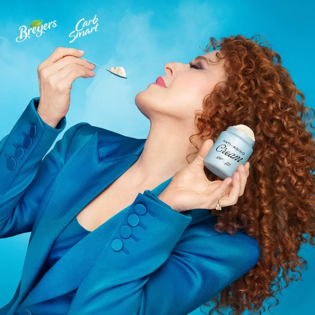 Bernadette Peters eats ice cream from a spoon, holding a container labeled "Anti-Aging Cream," in a light blue setting.
