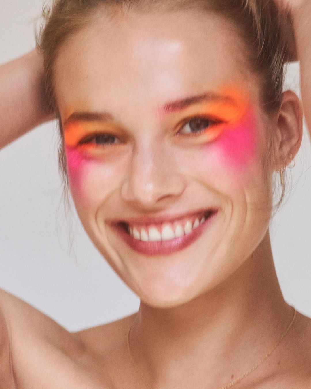 Close-up of a smiling woman with vibrant orange and pink makeup around her eyes, set against a neutral white backdrop.