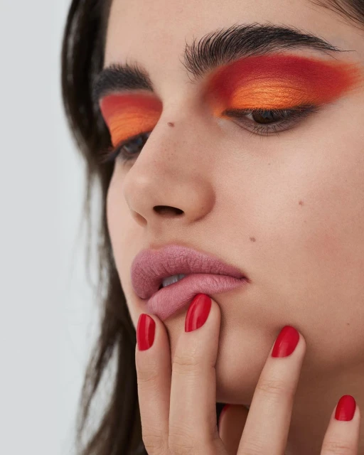 Close-up of a woman with bright orange and red eyeshadow and pink lipstick. Red manicured nails touch her face against a plain background.