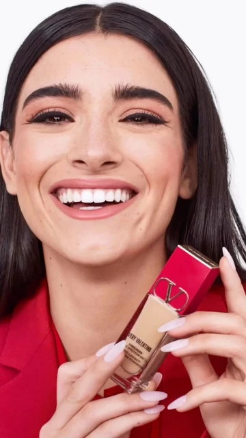 Woman smiling holding Valentino Very Valentino 24 Hour Wear liquid foundation makeup product in photo studio with white background.