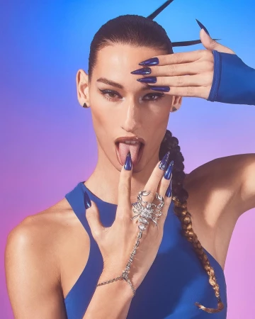 Woman with tongue out, blue nails, and a scorpion ring poses against a gradient background of blue and purple in a beauty style shot.