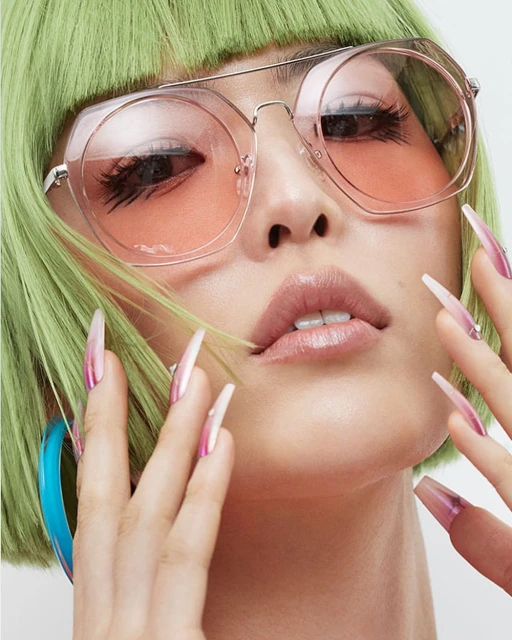 Close-up of a woman with green hair, pink sunglasses, blue hoop earring, and long, iridescent pink and white fingernails.