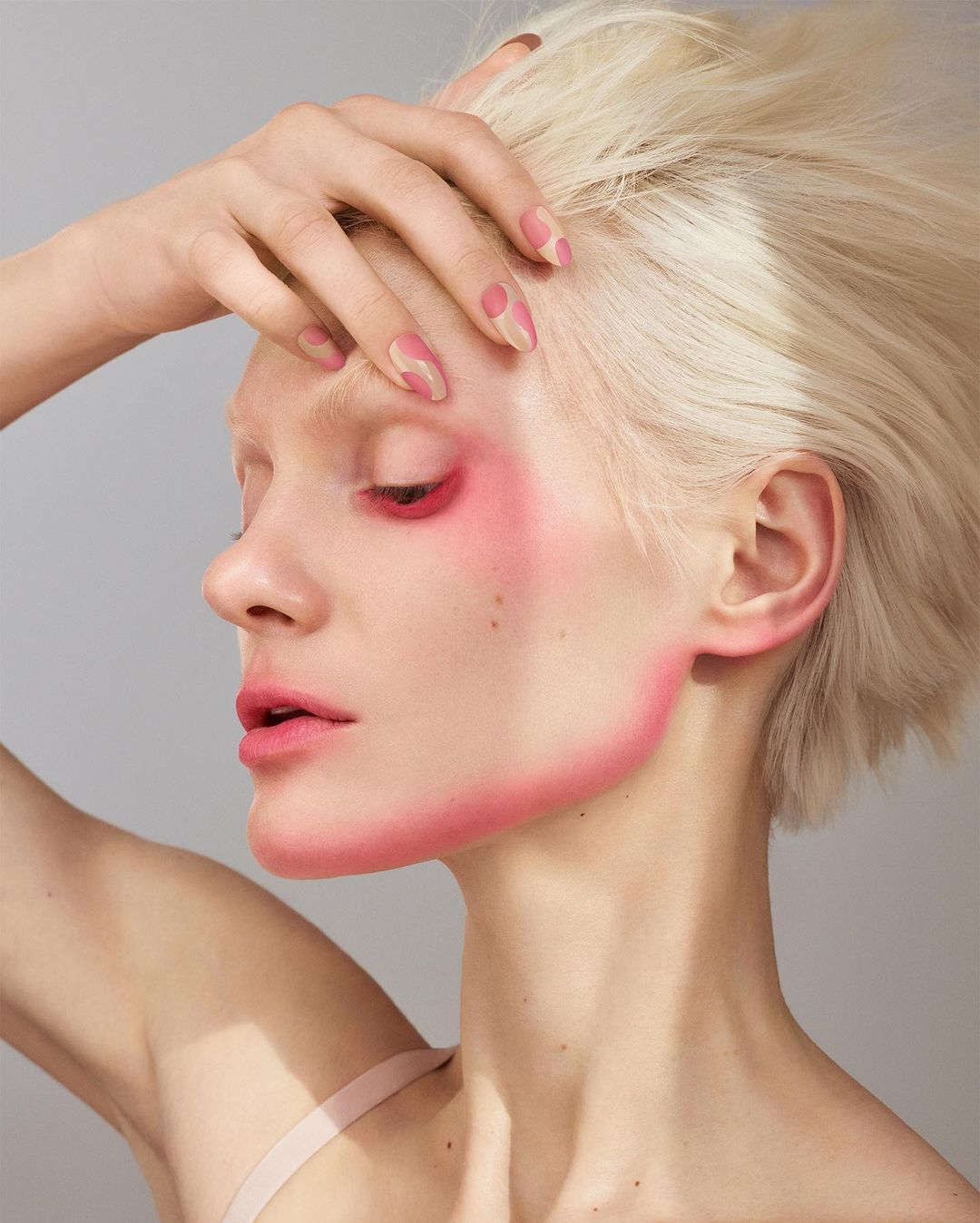 Adult female with pink face paint and hand on head poses in a beauty shot against a grey background.