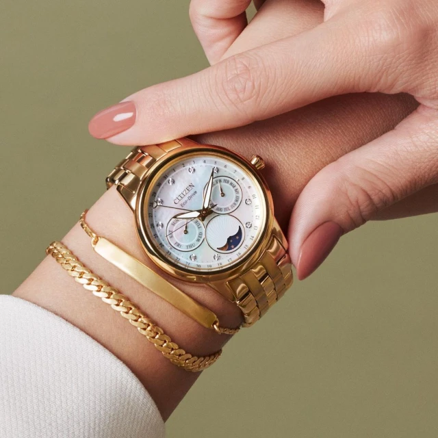 Close-up of a fair-skinned adult woman wearing a gold Citizen wristwatch and gold bracelets against an olive-green background.