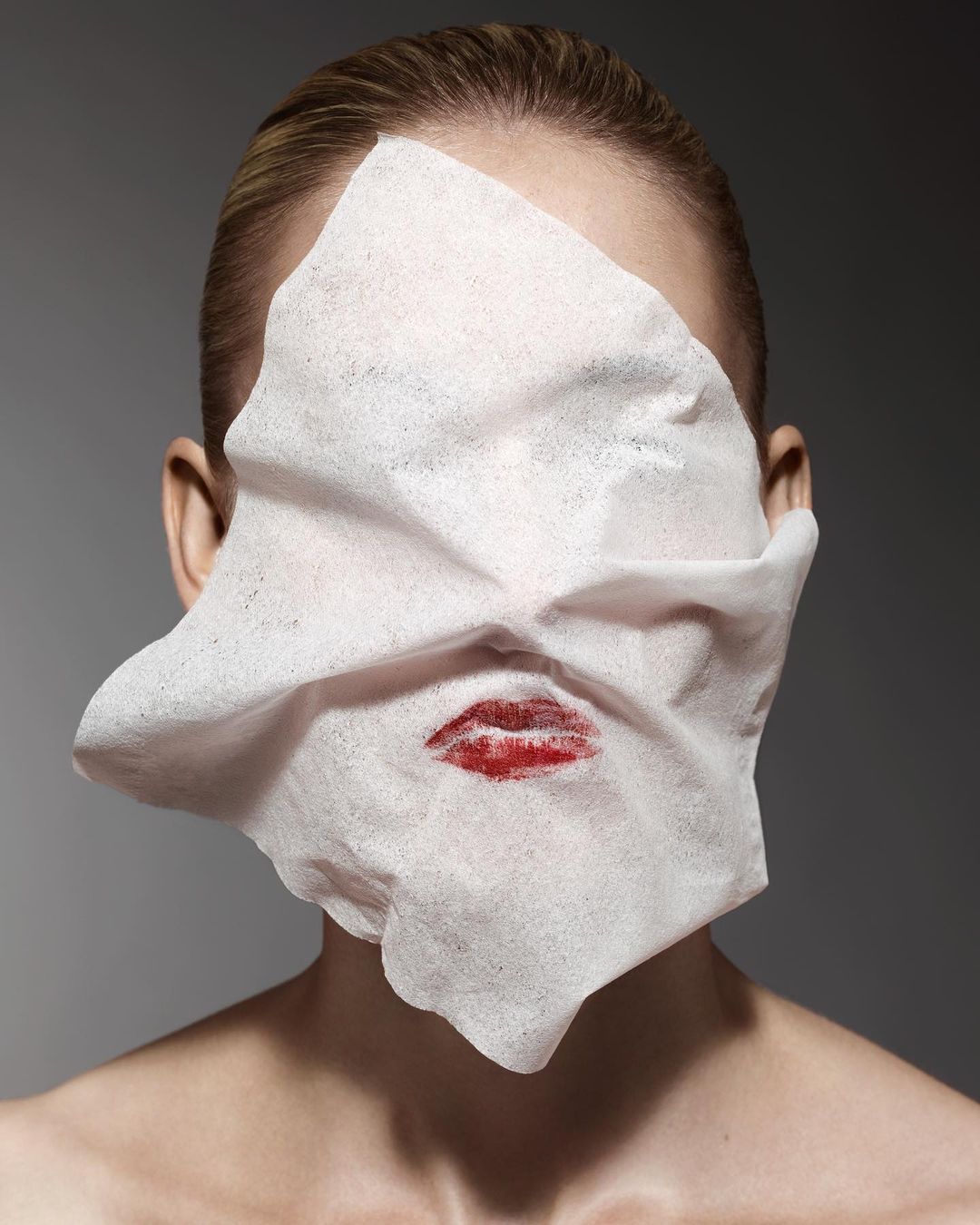 Portrait of a woman with lipstick kiss on a tissue covering her face against a grey background in a beauty-themed photograph.