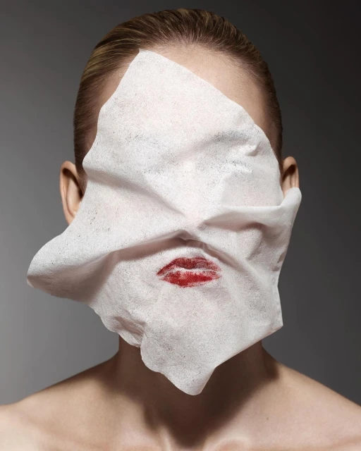 Portrait of a woman with lipstick kiss on a tissue covering her face against a grey background in a beauty-themed photograph.