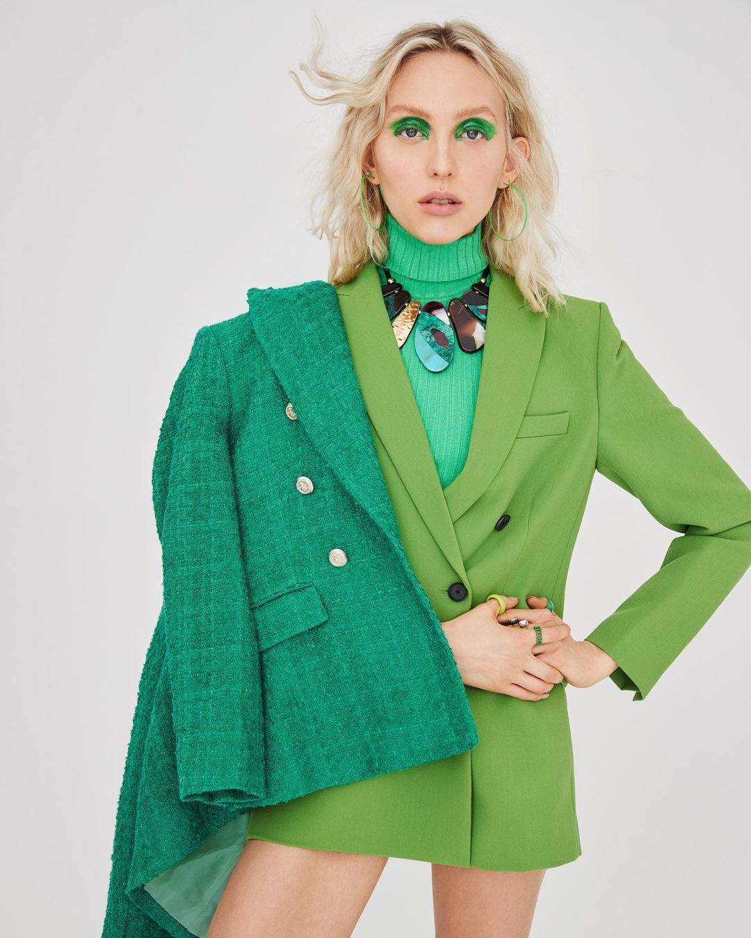 A woman in a green turtleneck and blazer, with bright green eyeshadow and large necklace, poses against a white backdrop in a studio shot.