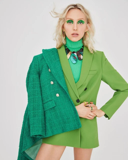 A woman in a green turtleneck and blazer, with bright green eyeshadow and large necklace, poses against a white backdrop in a studio shot.
