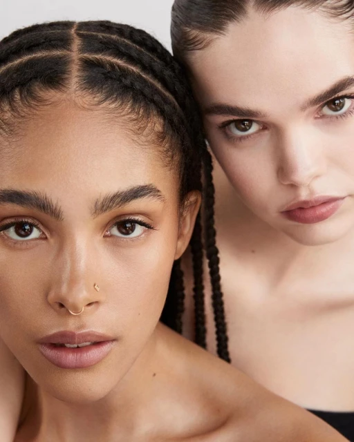 Close-up studio shot of two women with dark hair and neutral makeup posing together against a white backdrop.