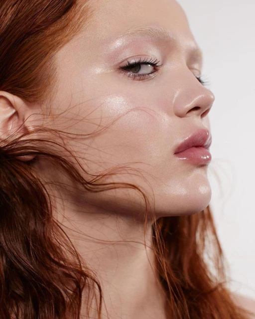 Close-up beauty shot of a redhead woman with fair skin and glossy lips, facing right with light makeup, set against a white backdrop.