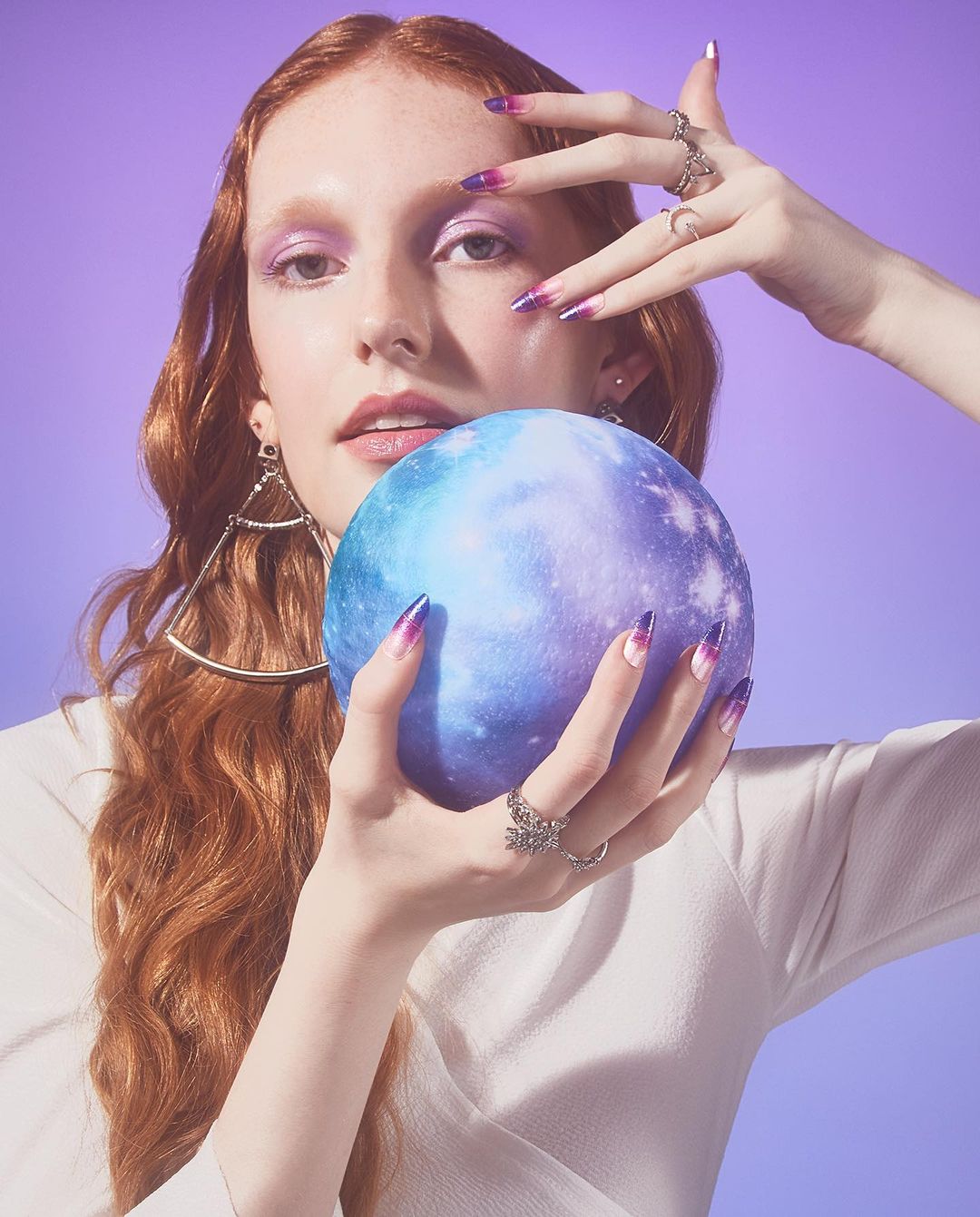 Red-haired woman with galaxy-themed makeup and nails holds a matching sphere in a beauty shot against a purple backdrop.