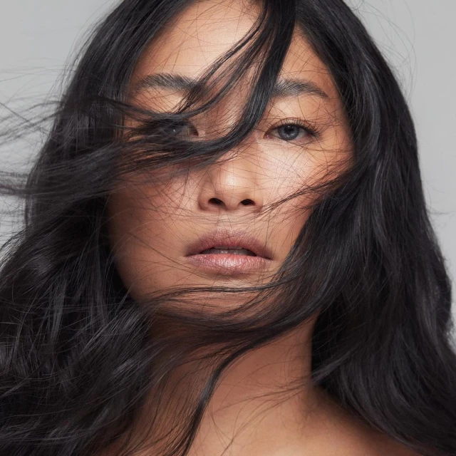 Close-up portrait of a young adult woman with fair skin and flowing black hair partially obscuring her face against a grey background.