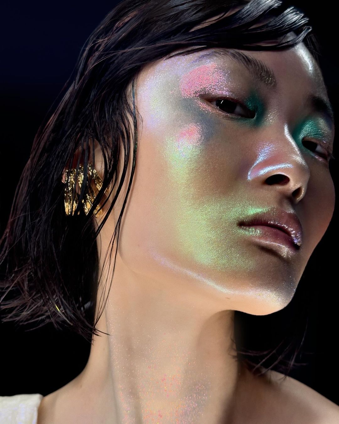 Close-up portrait of an Asian woman with pink and teal eye makeup, glossy lips, wet-look dark hair, and gold geometric earrings against a black background.