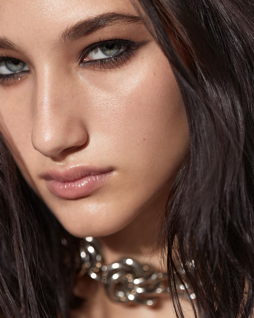 Close-up studio portrait of a young adult female with smokey eye makeup, dark hair, and a chunky silver chain necklace.
