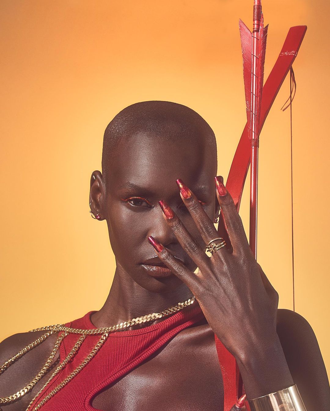 Elegant portrait of a dark-skinned woman with a shaved head and striking red nails holding a red bow and arrow against an orange background.