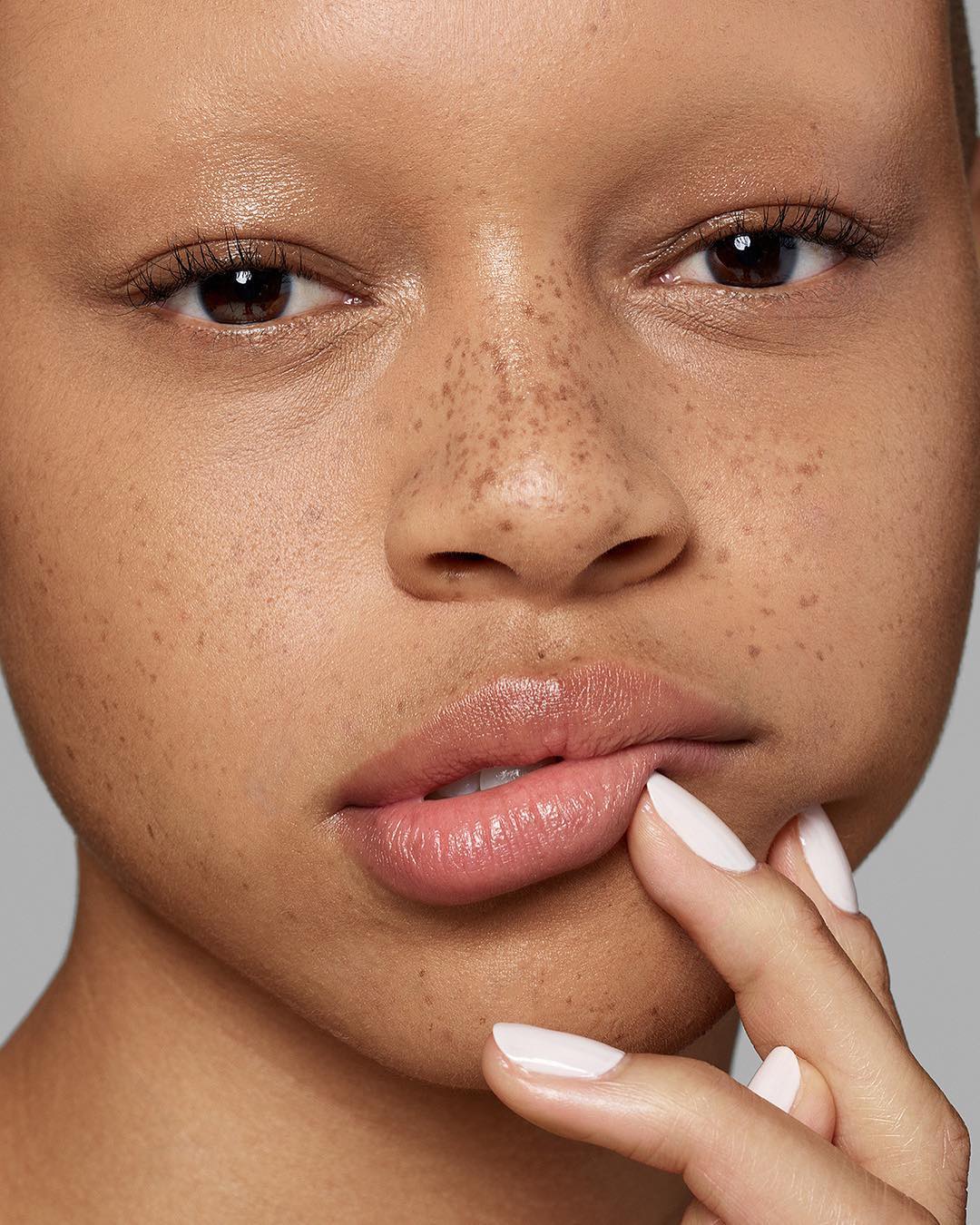 Close-up shot of a woman's face with fair skin and freckles, featuring her brown eyes, full lips, and a finger near her mouth.