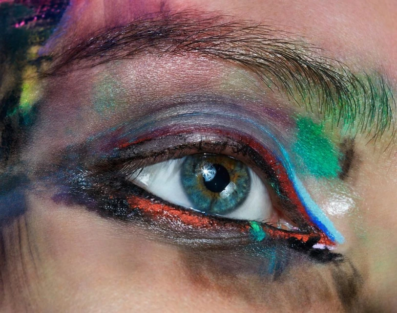 Close-up of a person's eye with vibrant makeup in shades of green, red, blue, and purple, creating an artistic and colorful effect.