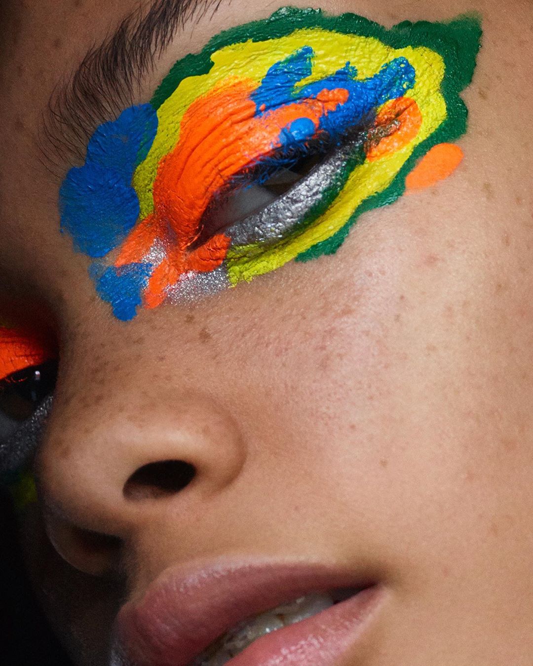 Eye-level close-up shot of a woman's face showing a vibrant, artistic eye makeup design featuring green, yellow, orange, and blue colors.